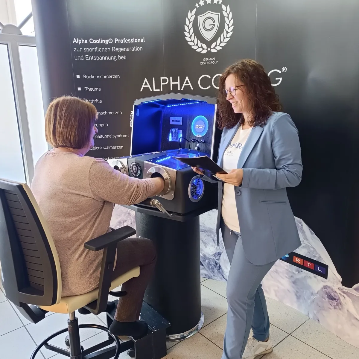 Margit Gartner im Gespräch mit einer Kundin, die am Alpha Cooling Gerät sitzt und die Hände eingelegt hat